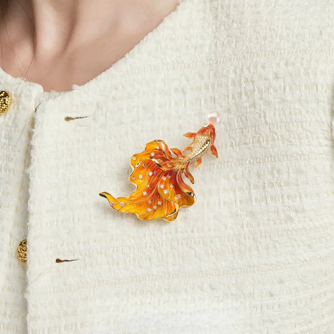 Aiko Tanaka Kasane brooch, an orange enamel goldfish with a pearl, pinned to a cream tweed jacket with gold buttons