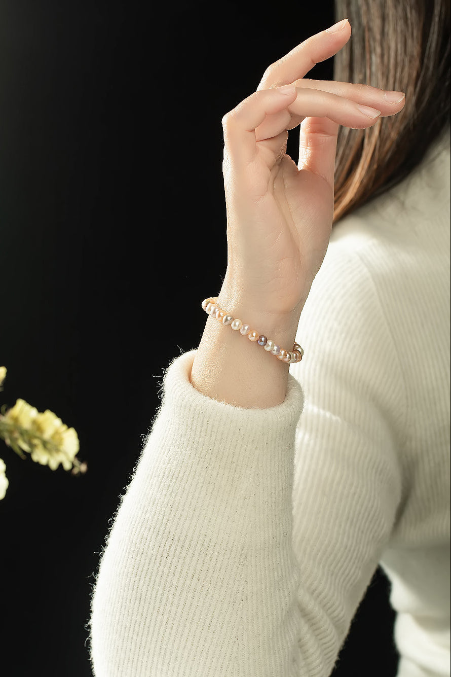 Raised hand in a cream sweater wearing the Aiko Tanaka Nagomi peach and white pearl bracelet against a black backdrop
