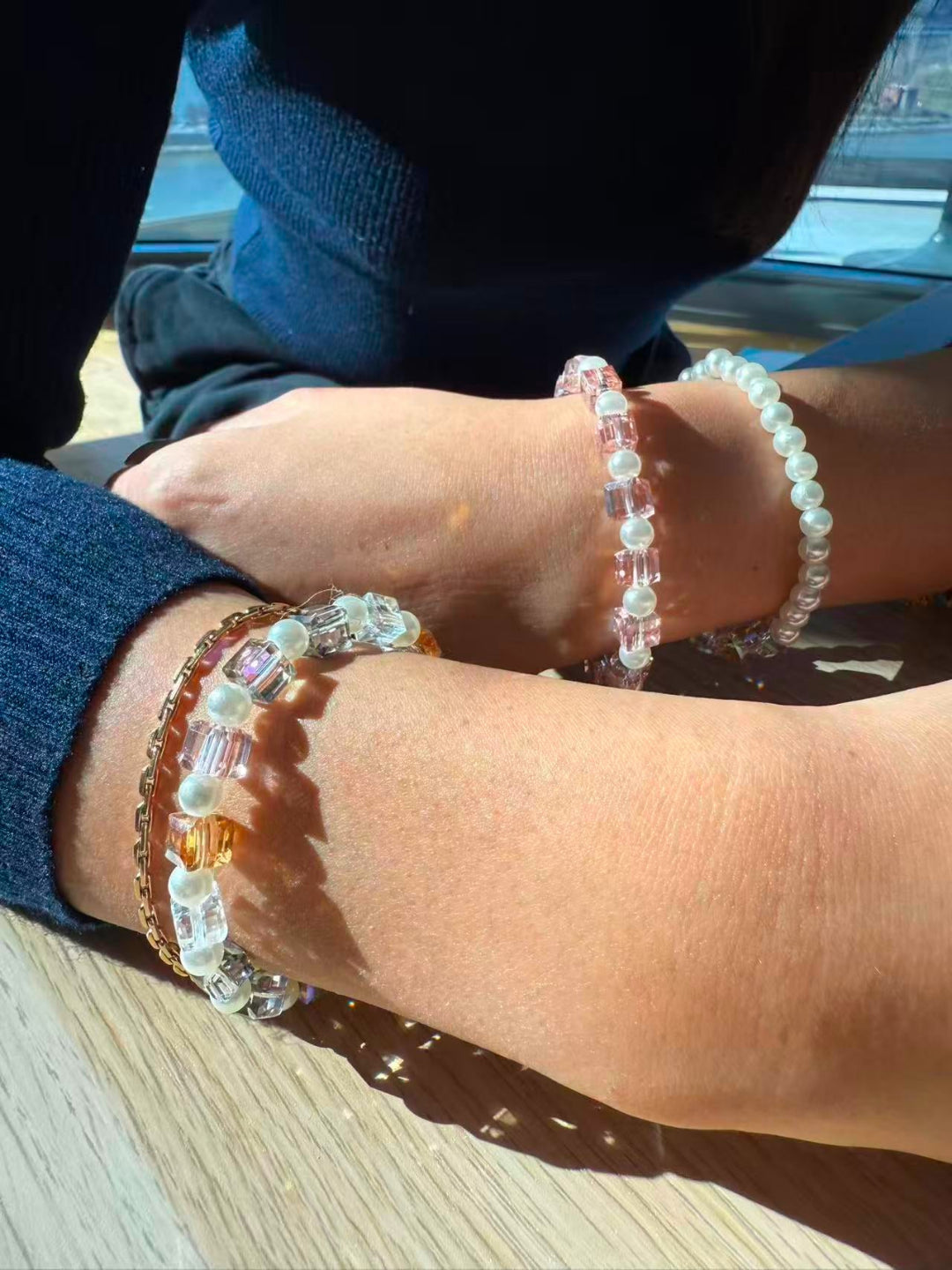 Two wrists wearing stacked Aiko Tanaka Kosai no Wa pearl and pastel crystal bracelets, resting on a sunny table