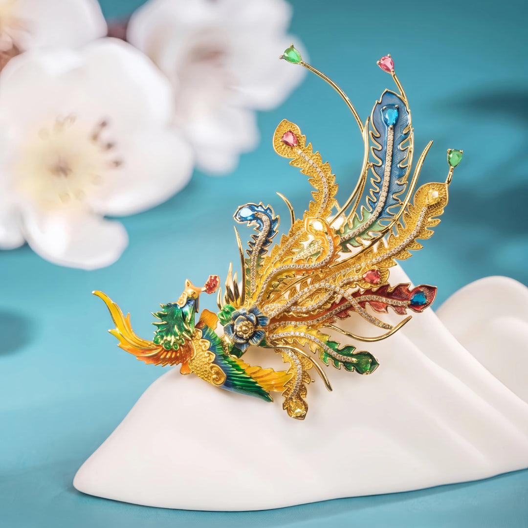 Aiko Tanaka Hoo no Kinka phoenix brooch in gold with colorful enamel plumes on a white stand beside cherry blossoms
