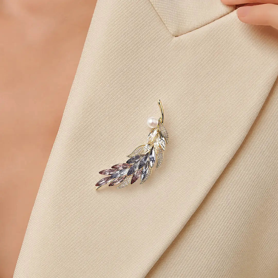 A gold-toned leaf-shaped brooch with a single pearl and rhinestone accents, pinned to a beige blazer.