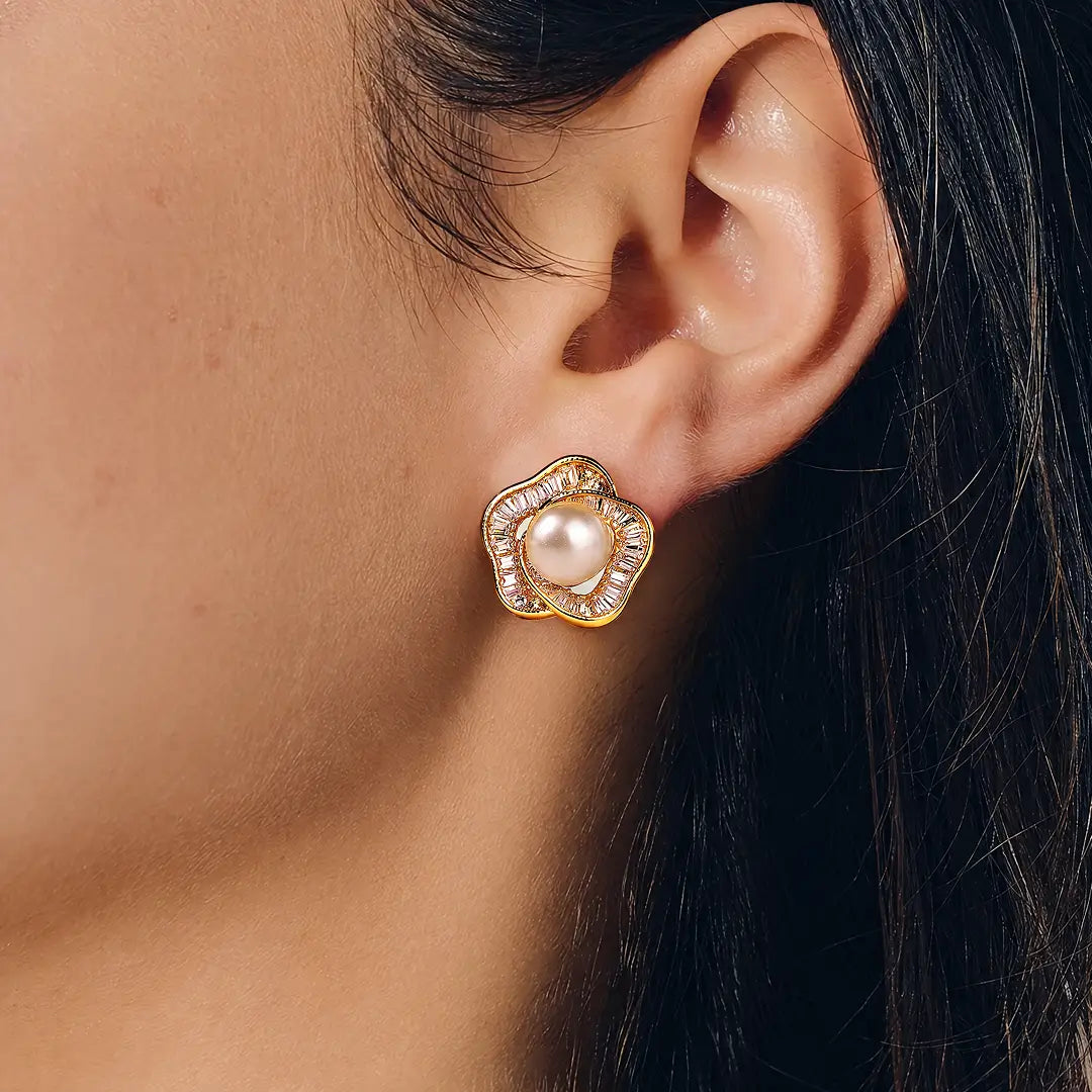 A close-up of a model's ear wearing a gold flower-shaped stud earring with a central white pearl and clear stones around it.