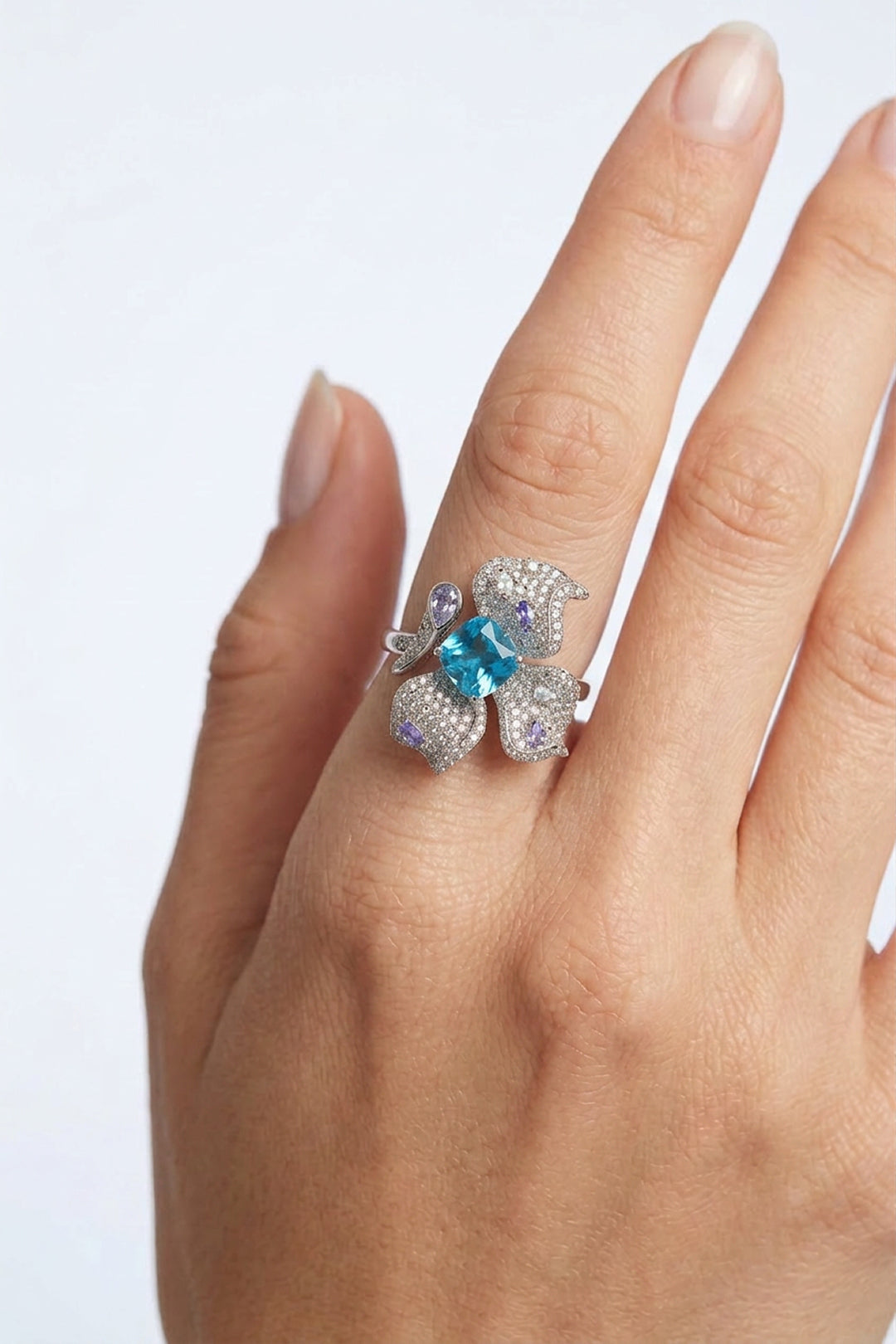 Hand wearing the Aiko Tanaka Mizu no Ran orchid ring with lilac crystal petals and an aqua blue stone, white backdrop