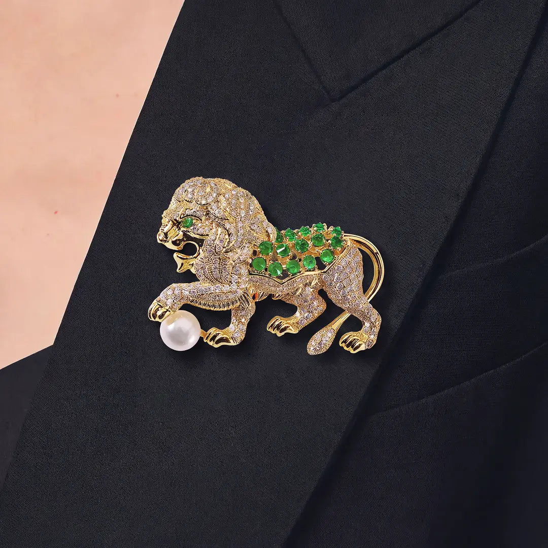 A gold lion-shaped brooch embellished with clear pavé stones, green gemstones on its back, and a white pearl bead at its mouth, pinned to a black blazer.