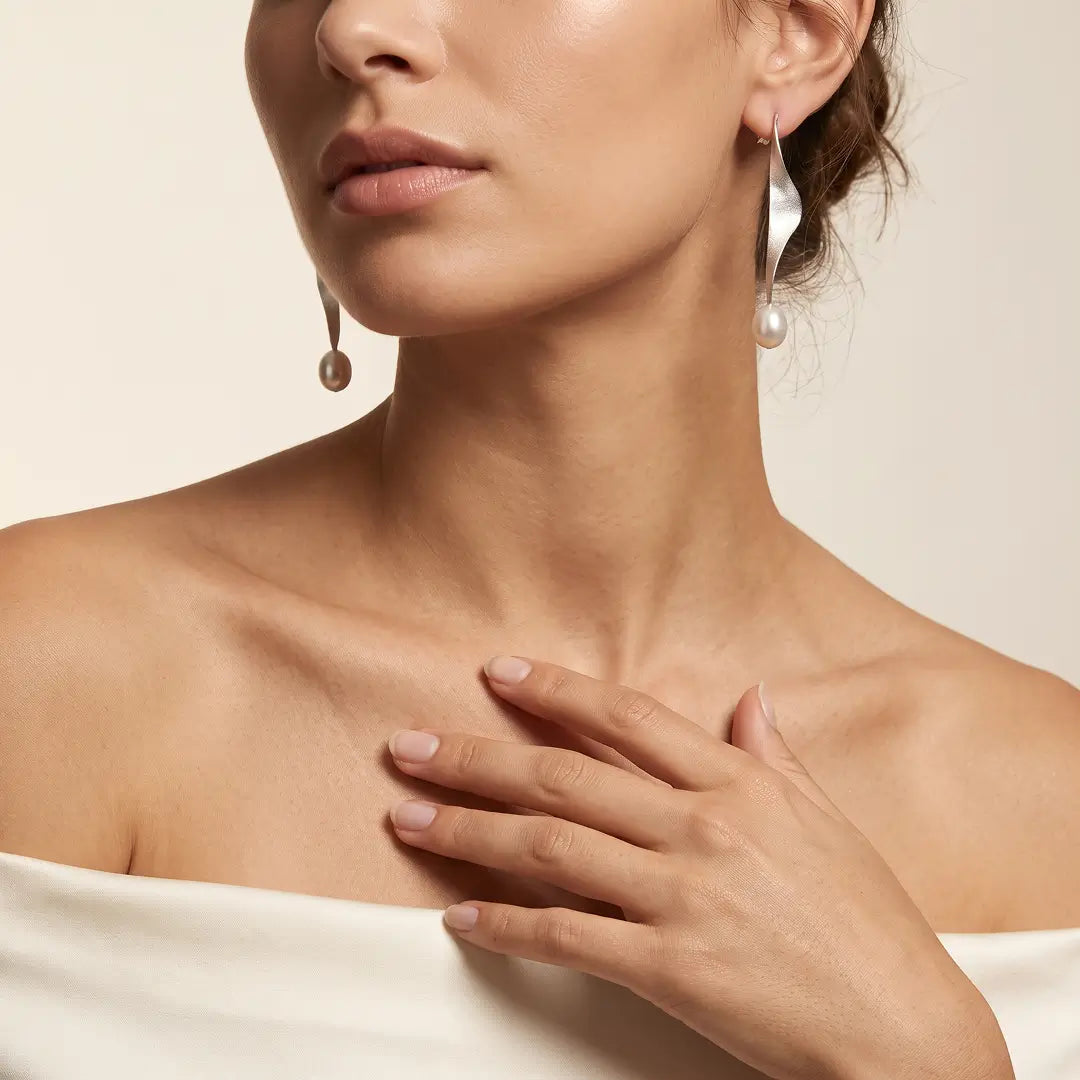 Aiko Tanaka Shizuku earrings, a twisted silver ribbon drop ending in a pearl, worn by a model in an off-shoulder top